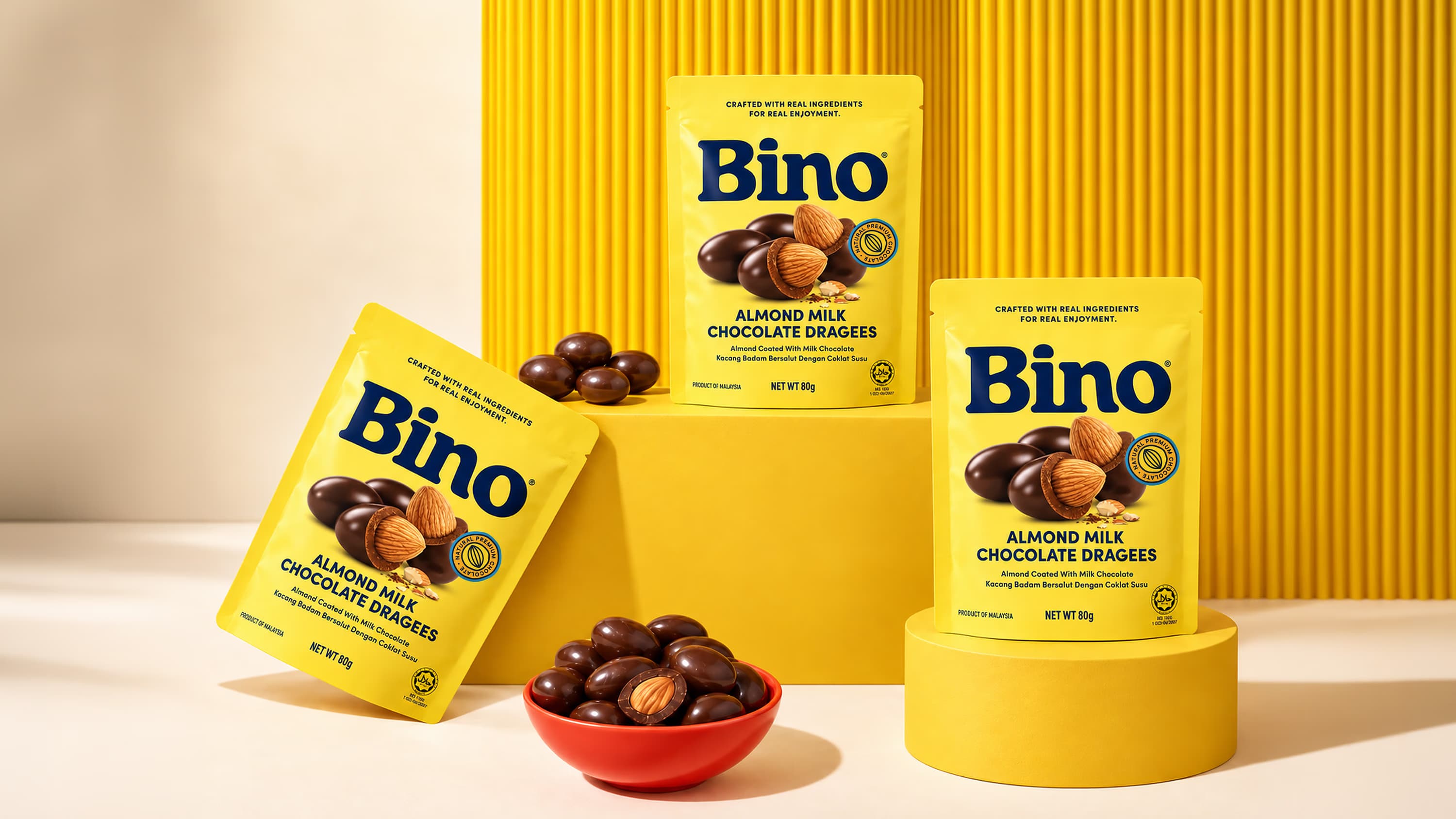 Three yellow packs of BINO Almond Milk Chocolate Dragees arranged against a yellow background.